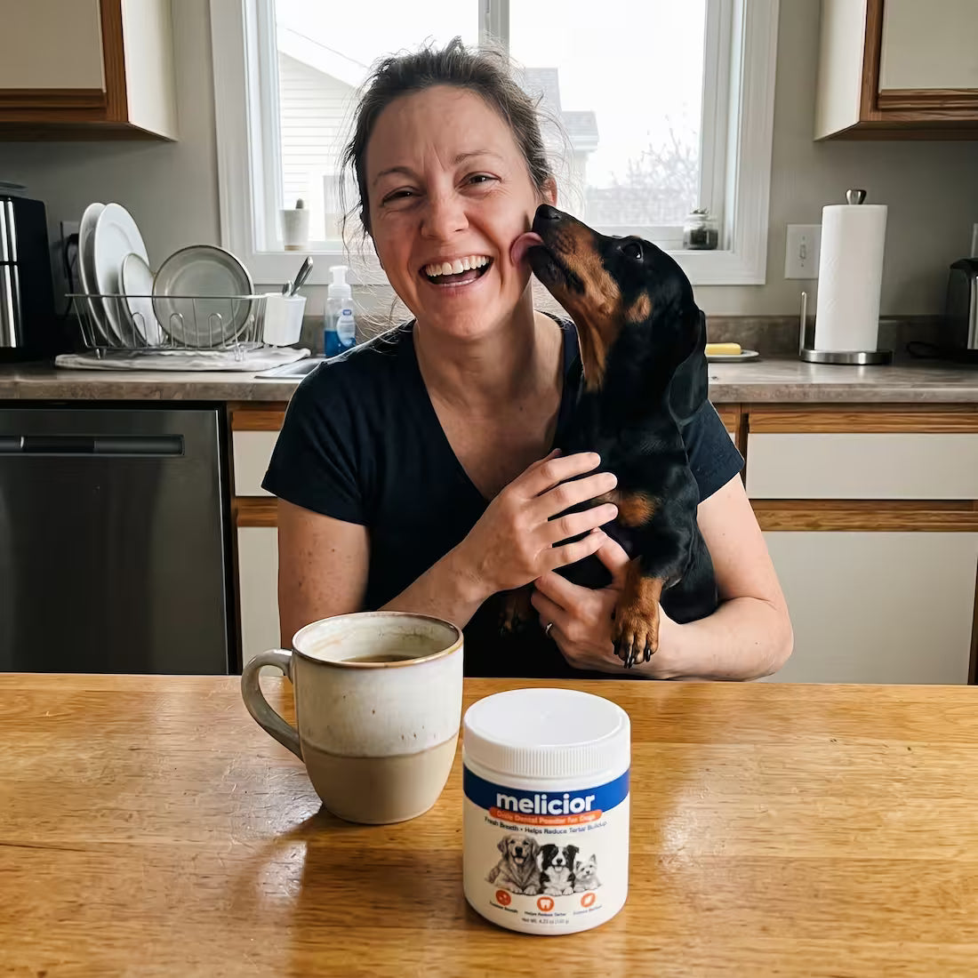 A woman laughs as her dachshund licks her cheek, a jar of Melicior dental powder on the kitchen table